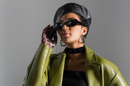 Portrait of trendy african american woman in sunglasses and leather coat isolated on grey 