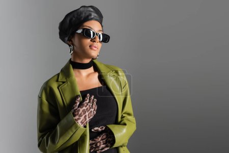 Fashionable african american woman in sunglasses and green coat standing isolated on grey 