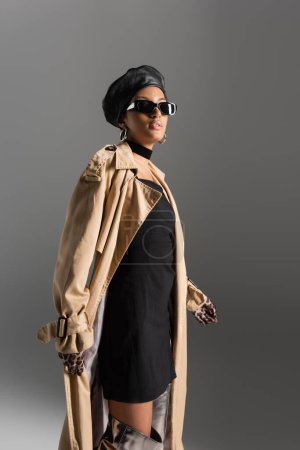 Stilvolle afrikanisch-amerikanische Frau in Sonnenbrille und Trenchcoat isoliert auf grau 