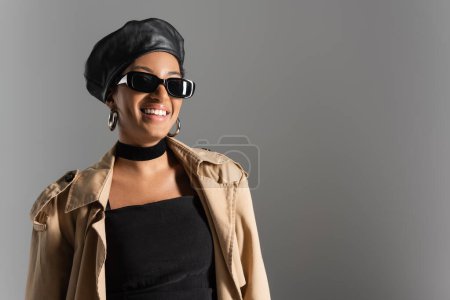 Téléchargez les photos : Femme afro-américaine souriante en béret et trench coat isolés sur gris - en image libre de droit