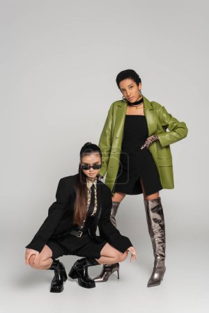 Junge multiethnische Freundinnen in stylischer Kleidung posieren auf grauem Hintergrund