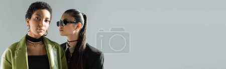 Portrait of stylish african american model standing near asian friend isolated on grey, banner 