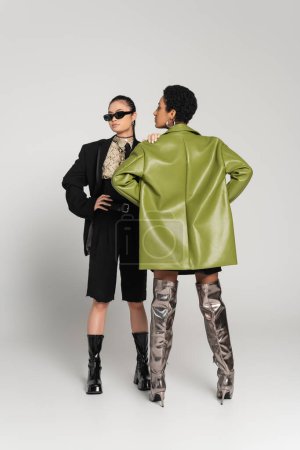 Full length of stylish asian model posing near african american friend in leather coat on grey background