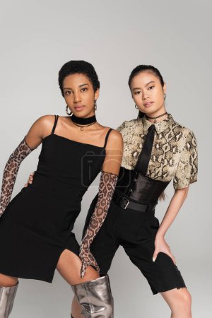 Trendy multiethnic women looking at camera while posing in clothes with animals prints isolated on grey 