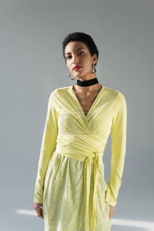 Trendige Afroamerikanerin im gelben Outfit blickt in die Kamera auf grauem Hintergrund
