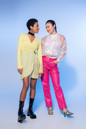 Smiling interracial women in bright outfit looking at each other on blue background 