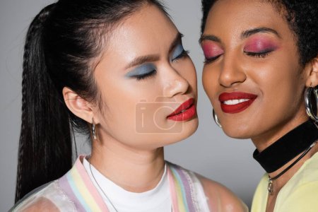 Asian woman with colorful makeup closing eyes near smiling african american friend on grey background 