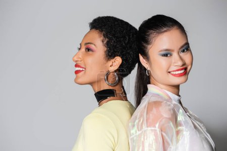 Pretty smiling interracial models with red lips standing back to back on grey background 