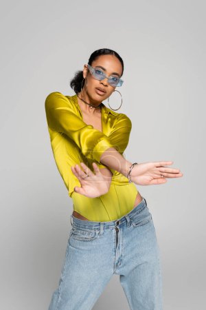 african american woman in blue sunglasses and stylish outfit posing isolated on grey  