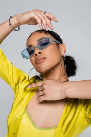 pretty african american woman in blue sunglasses and stylish outfit gesturing isolated on grey  