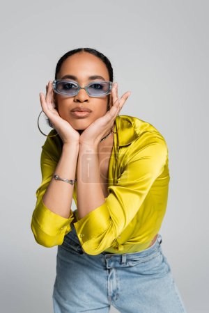 pretty african american woman in blue sunglasses touching face and looking at camera isolated on grey