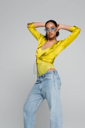 Téléchargez les photos : Brunette modèle afro-américain en lunettes de soleil bleues posant et regardant la caméra isolée sur gris - en image libre de droit
