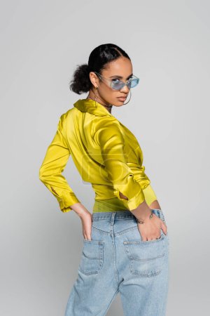 young african american model in blue sunglasses and trendy clothes posing with hands in back pockets isolated on grey 