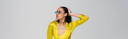 pretty african american woman in stylish sunglasses touching head while posing isolated on grey, banner 