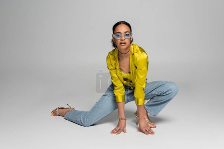 in voller Länge von stylischem afrikanisch-amerikanischem Model in blauer Sonnenbrille posiert auf Knien vor grauem Hintergrund 