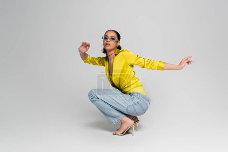 full length of stylish african american woman in high heels and trendy outfit sitting on haunches on grey 