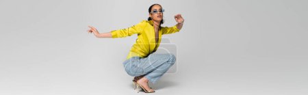 full length of stylish african american model in high heels and trendy outfit sitting on haunches on grey, banner  