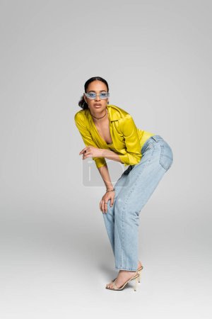 full length of stylish african american woman in trendy outfit and high heels posing on grey 