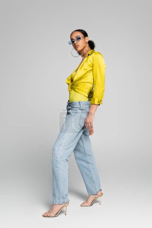 Téléchargez les photos : Toute la longueur du modèle afro-américain élégant en lunettes de soleil à la mode et talons hauts posant sur le gris - en image libre de droit