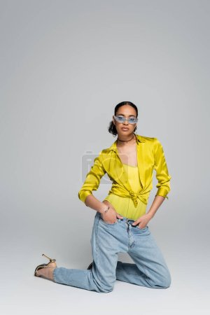 Photo for Full length of pretty african american woman standing on knees while posing with hands in pockets on grey - Royalty Free Image
