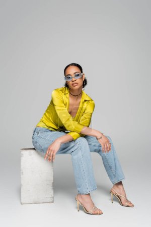 in voller Länge stilvolle afrikanisch-amerikanische Frau in trendiger Sonnenbrille und Outfit sitzt auf Betonwürfel, während sie auf grau posiert 