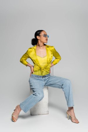 Afrikanisches amerikanisches Model in voller Länge mit stylischer Sonnenbrille auf Betonwürfel sitzend, während sie mit Händen auf Taille auf grau posiert 