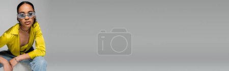 Téléchargez les photos : Jeune modèle afro-américain en lunettes de soleil élégantes regardant la caméra sur fond gris, bannière - en image libre de droit