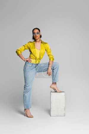 full length of confident african american model stepping on concrete cube while posing with hand on hip on grey 