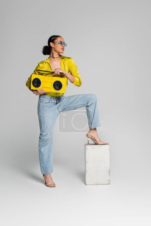 brünettes afrikanisch-amerikanisches Model tritt auf Betonwürfel und hält gelbe Boombox auf grau 