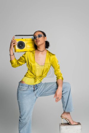 brunette african american woman in blue sunglasses stepping on concrete cube while holding yellow boombox on grey 