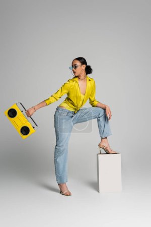 full length of brunette african american woman in sunglasses stepping on concrete cube while holding retro boombox on grey 