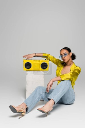 full length of brunette african american woman in sunglasses sitting near concrete cube and retro boombox on grey 