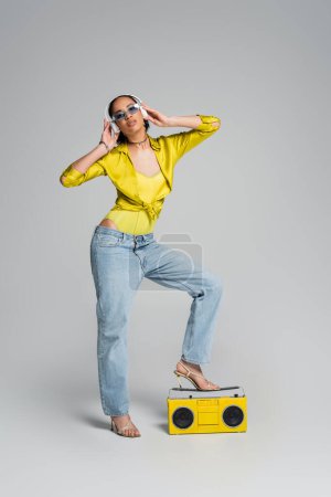 full length of african american woman in wireless headphones enjoying music near yellow boombox on grey 