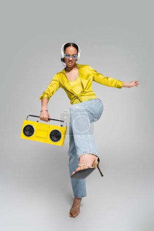 full length of emotional african american model in wireless headphones holding yellow boombox and posing on grey 