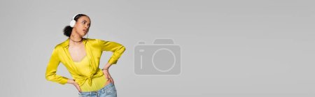 stylish african american model listening music in wireless headphones and posing with hands on hips isolated on grey, banner 
