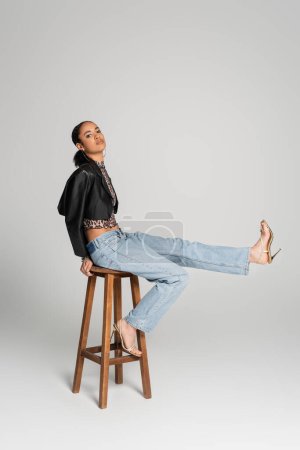 Téléchargez les photos : Toute la longueur du modèle afro-américain à la mode en veste recadrée et sandales à talons hauts assis sur un tabouret haut sur gris - en image libre de droit