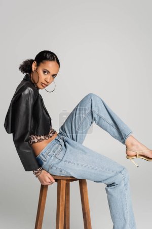 stylish african american woman in cropped jacket and jeans sitting on wooden high chair isolated on grey
