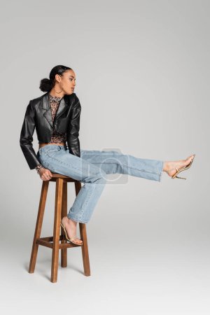 full length of young african american model in cropped jacket and jeans sitting on high stool on grey