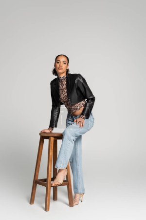 Téléchargez les photos : Toute la longueur du modèle afro-américain élégant en veste recadrée et jeans debout près de chaise haute en bois sur gris - en image libre de droit