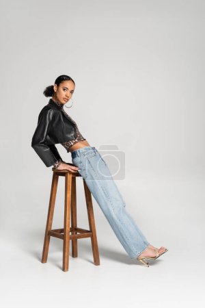 Téléchargez les photos : Pleine longueur de femme afro-américaine à la mode en veste et jeans cropped appuyé sur tabouret haut en bois sur gris - en image libre de droit