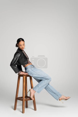 Téléchargez les photos : Toute la longueur du modèle afro-américain élégant en veste et jeans cropped appuyé sur chaise haute en bois sur gris - en image libre de droit