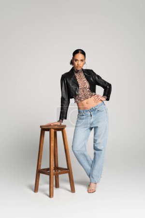 Photo for Full length of stylish african american woman in cropped jacket and jeans posing with hand on hip near high chair on grey - Royalty Free Image