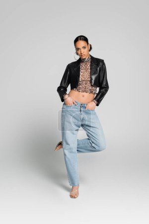 full length of african american model in cropped jacket posing with hands in pockets while standing on one leg on grey