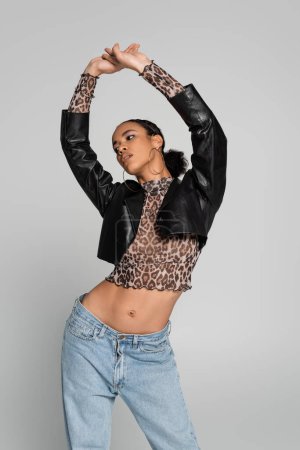 fashionable african american model in cropped jacket and jeans posing with raised hands isolated on grey