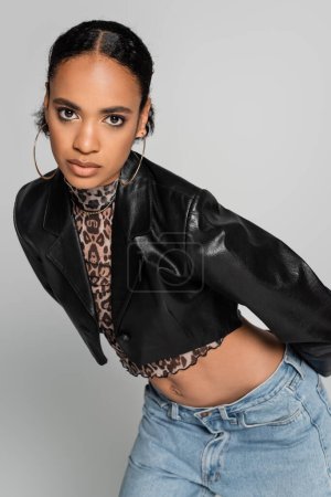 fashionable african american model in cropped jacket and hoop earrings posing isolated on grey