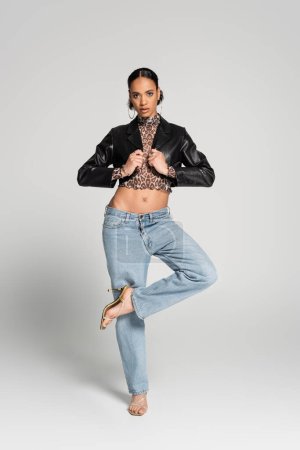full length of stylish african american model in cropped jacket and jeans posing on one led while standing on grey