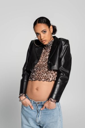 fashionable african american woman in black cropped jacket and top with animal print posing with hands in pockets isolated on grey 
