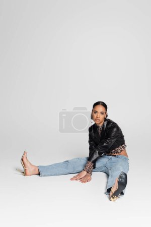 full length of stylish african american woman in high heeled sandals and trendy outfit sitting on grey 