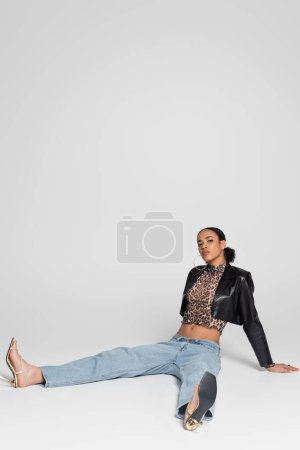 longitud completa de la joven afroamericana mujer en sandalias de tacón alto y traje de moda sentado en gris 