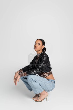 Photo for Full length of stylish african american woman in high heeled sandals and trendy outfit sitting on haunches on grey - Royalty Free Image